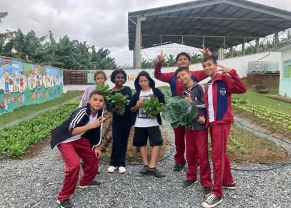 Foto de diversos alunos posando com algumas verduras nas mãos e uma horta ao fundo