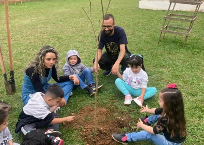 Crianças sentadas na grama ao redor de uma árvore sendo plantada